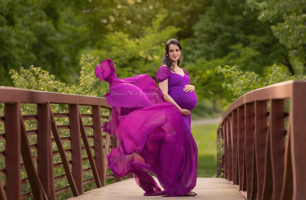 Pregnant woman in vibrant purple maternity dress standing on a bridge outdoors, celebrating motherhood.