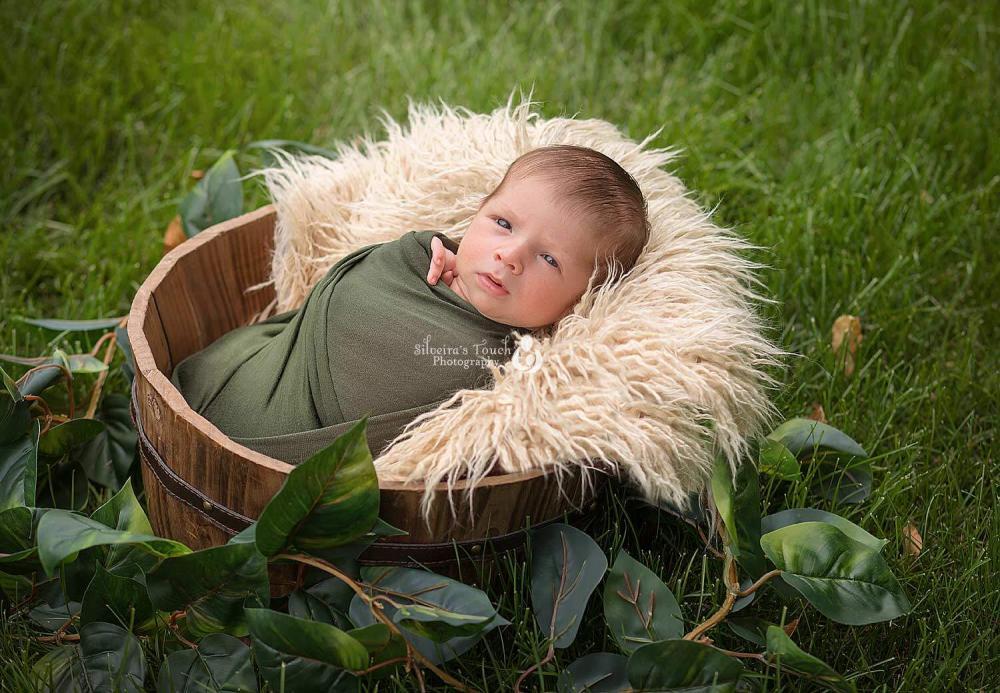 Baby lying in a wooden bucket with a fluffy blanket outdoors, surrounded by green grass and leaves, captured during a professional baby and maternity photography session in NJ.